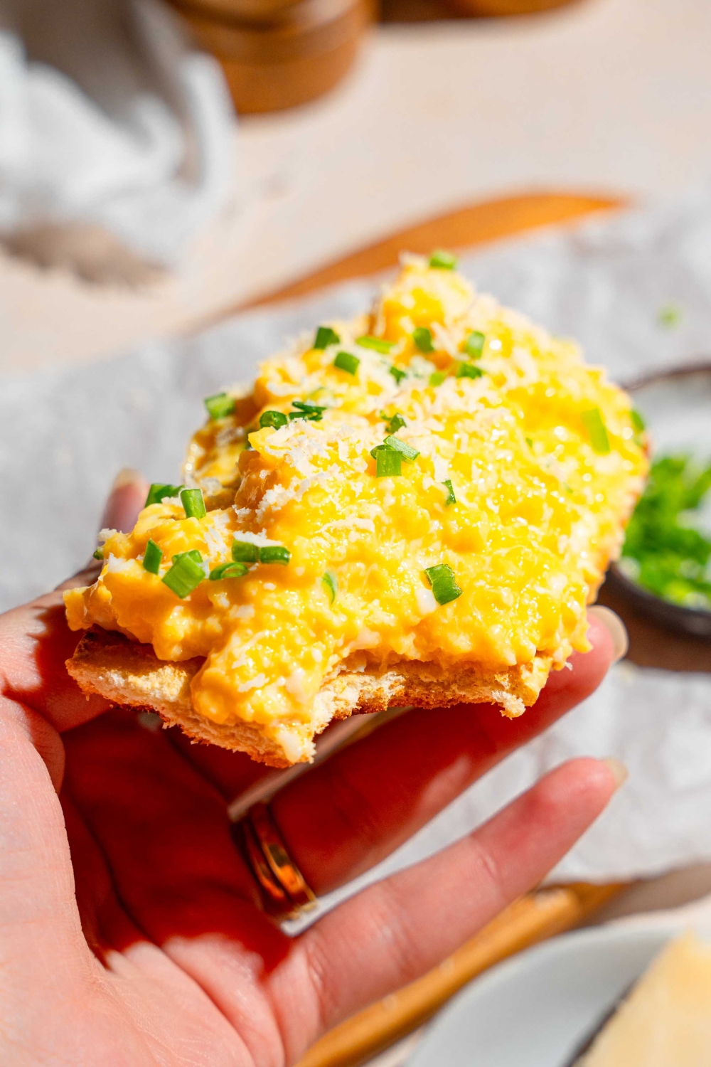 A close up of a hand holding a slice of thin toast topped with Gordon Ramsay scrambled eggs garnished with grated cheese and chives. There is a wooden board lined with parchment paper with a small bowl of garnishes blurred in the background.