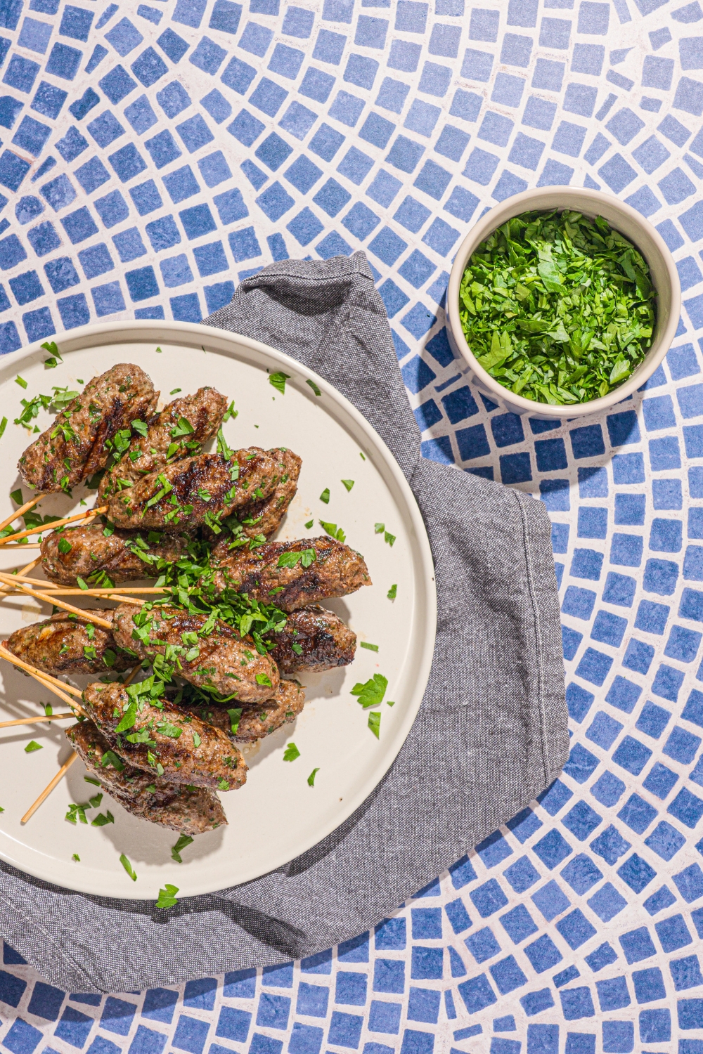 A white plate with a pile of Kafta kabobs garnished with fresh parsley. The plate is on a tiled counter with a gray cloth napkin.