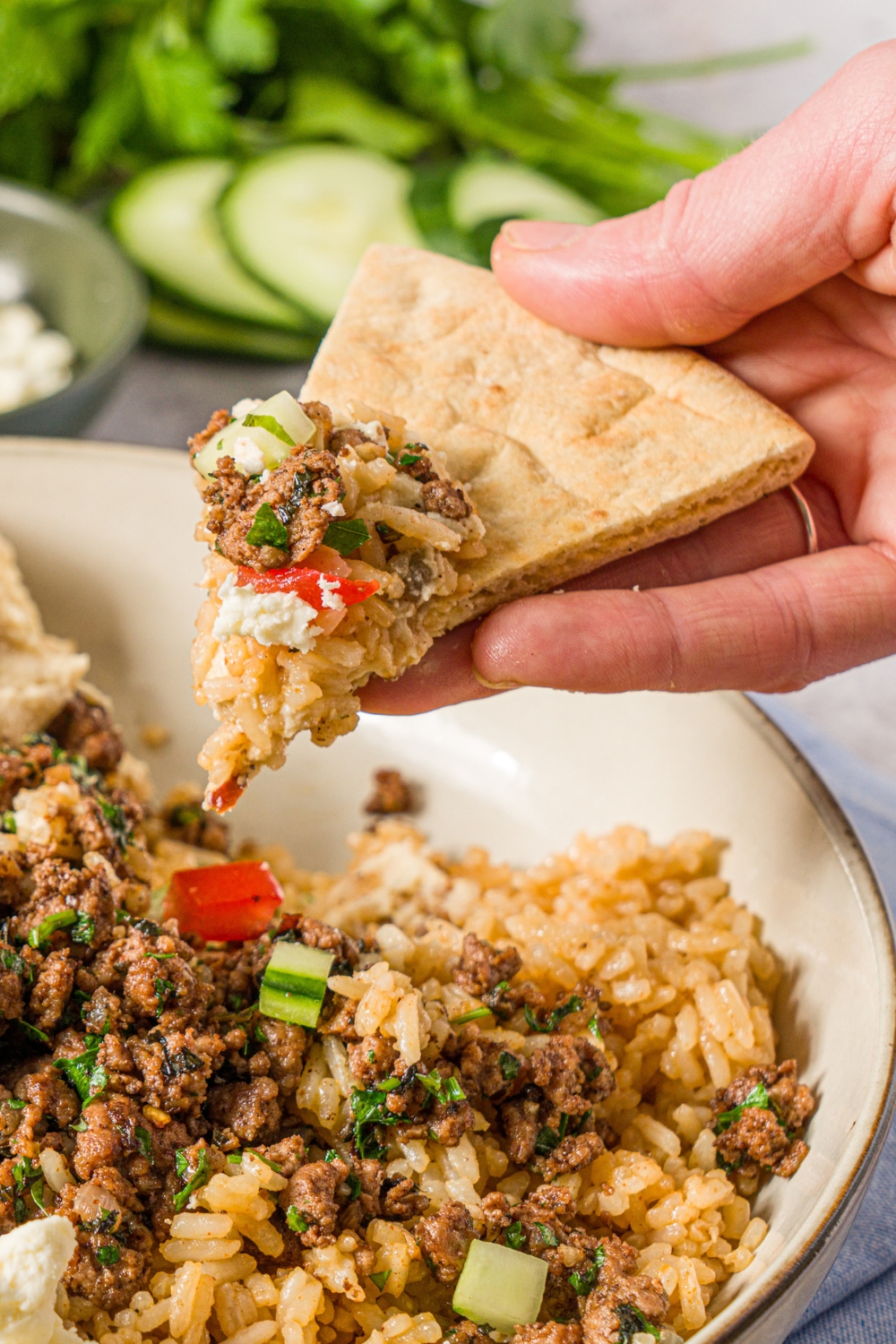 A hand with a slice of pita dipped into a Mediterranean rice bowl with brown rice, seasoned ground lamb, tomatoes, cucumbers, and feta cheese.