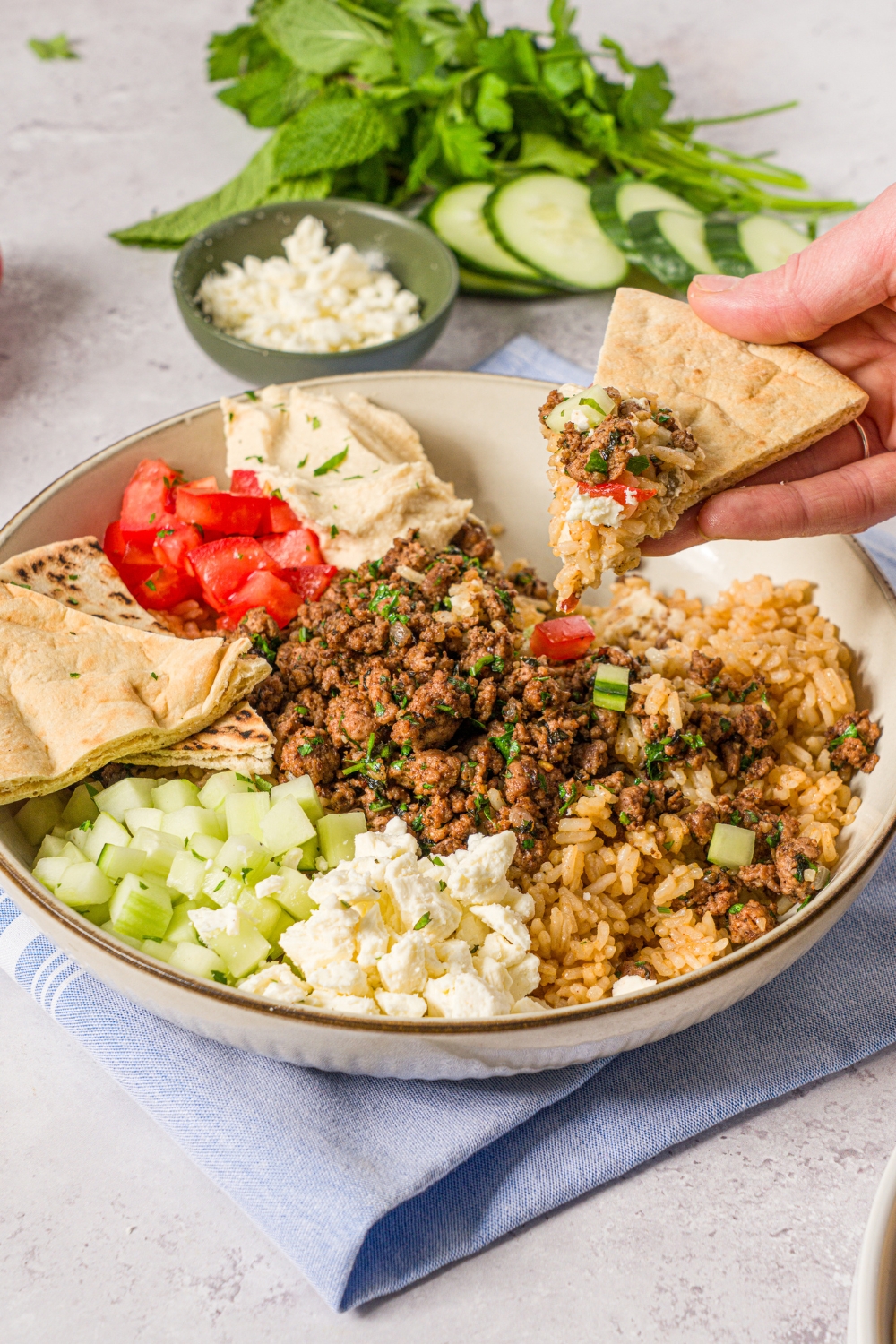 A hand with a slice of pita dipped into a Mediterranean rice bowl with brown rice, seasoned ground lamb, tomatoes, cucumbers, and feta cheese. The bowl is on a white counter with a blue cloth napkin.