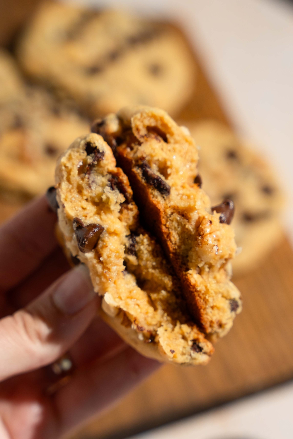 A close up of a hand folding a Neiman Marcus chocolate chip cookie in half. There is a wooden board with cookies blurred in the background.