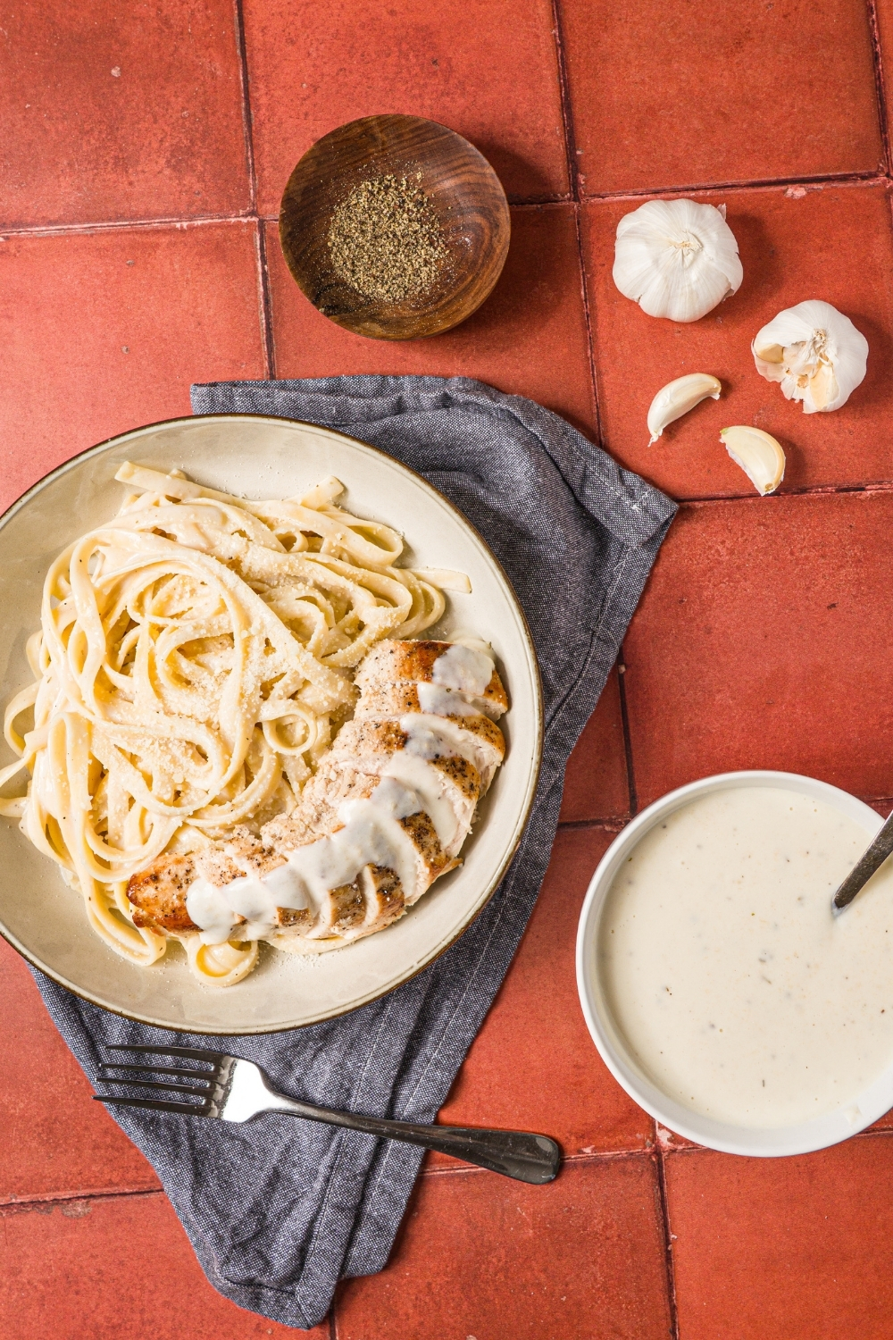 A bowl of chicken alfredo served with fettuccine pasta. The bowl is on a brick counter with a blue napkin and bowl of alfredo sauce.