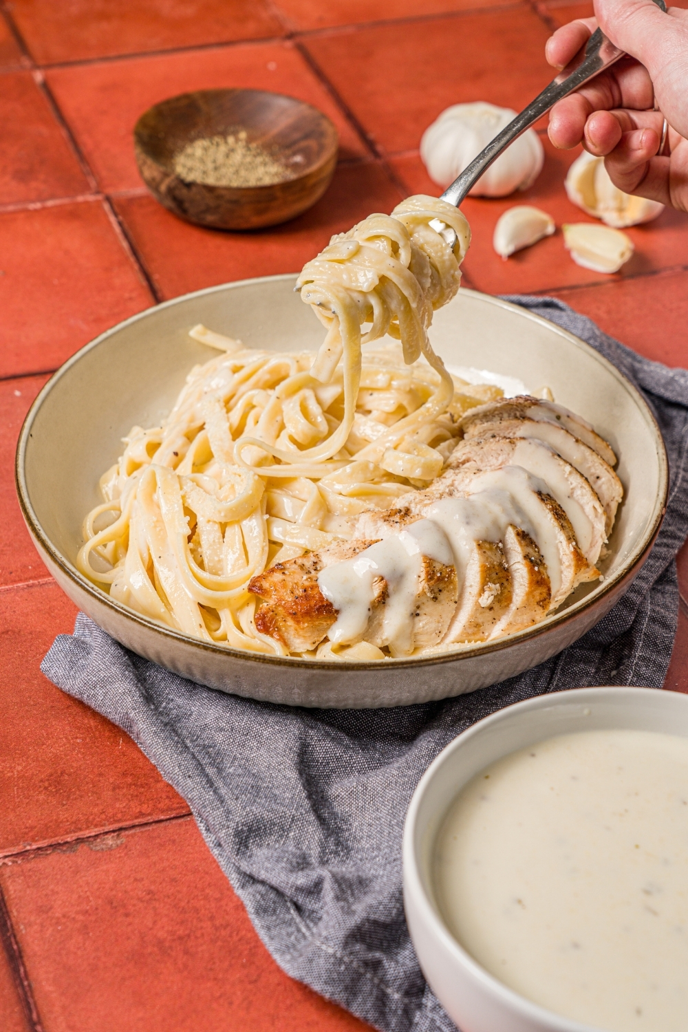 A bowl of chicken alfredo with a fork taking a bite of the fettuccine. The bowl is on a brick counter with a bowl of alfredo sauce.