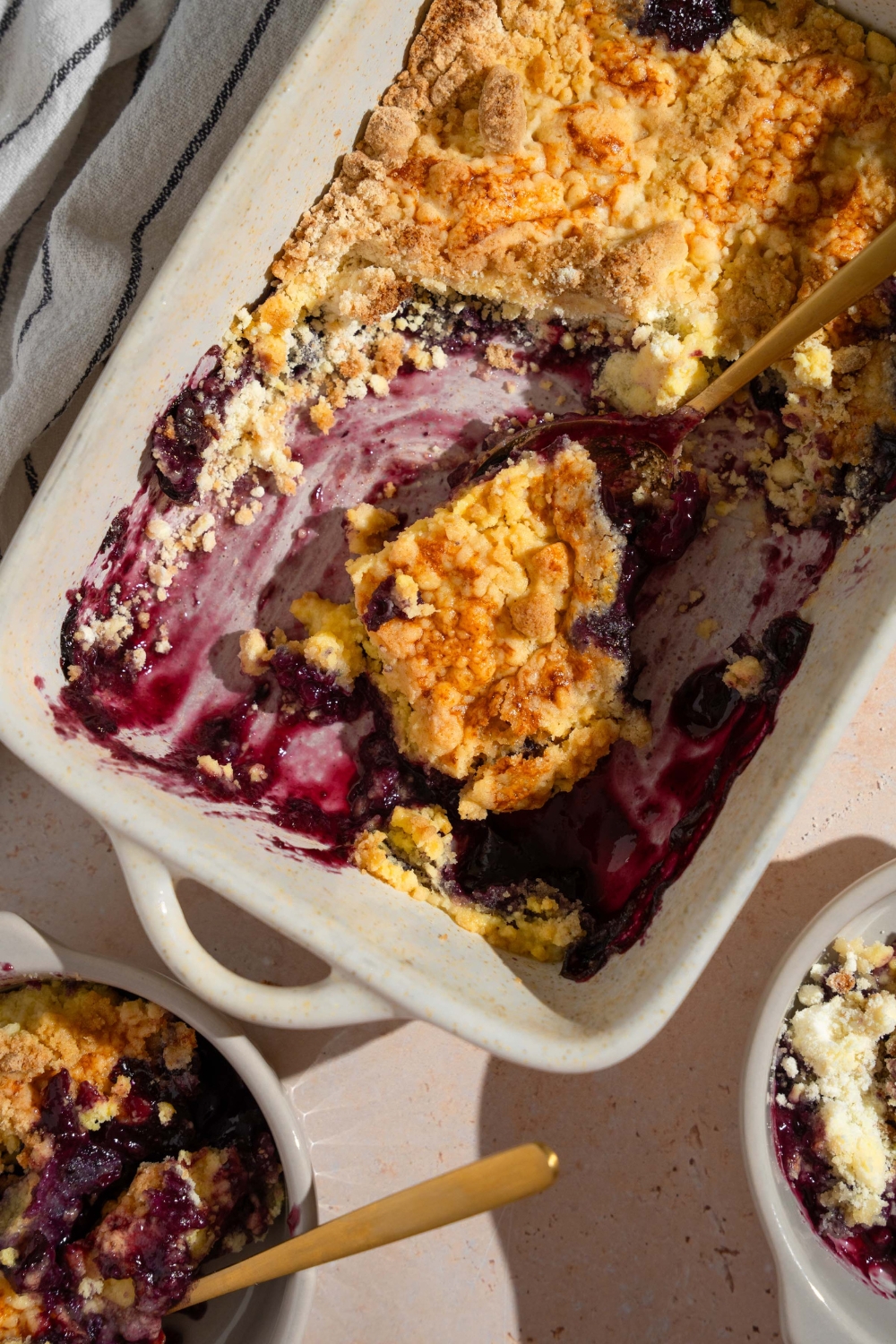 A baking dish with baked blueberry dump cake with a spoon taking a slice. The dish is on a tan counter with a bowl of dump cake.