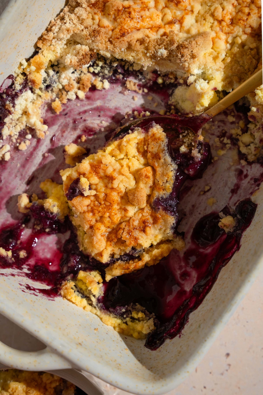 A baking dish with baked blueberry dump cake with a spoon taking a slice. The dish is on a tan counter with a bowl of dump cake.