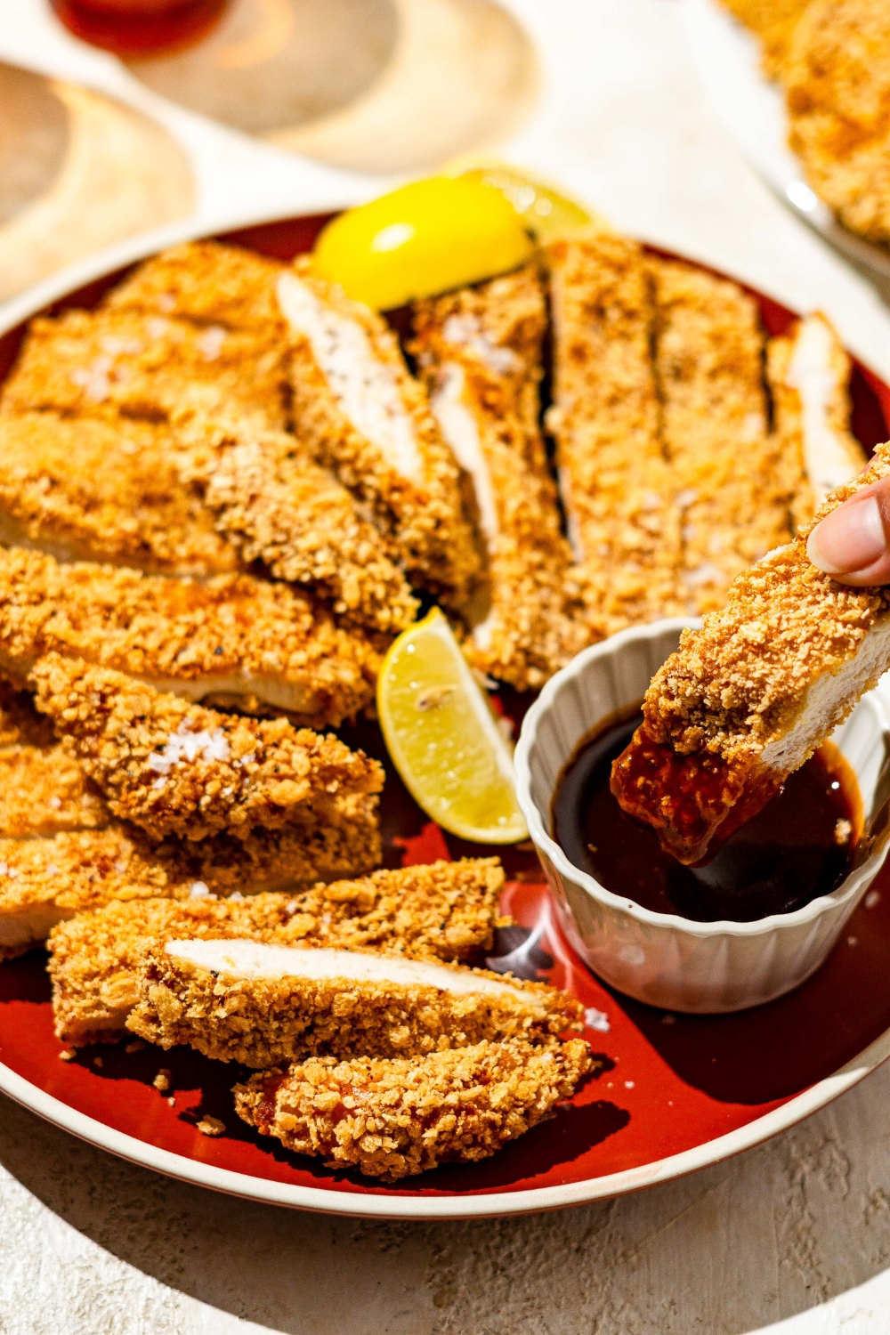 A plate of crispy chicken Katsu sliced in strips and served with lemon wedges and a side ramekin of sauce. A hand is dipping a chicken strip into the sauce. The plate is on a tan counter.