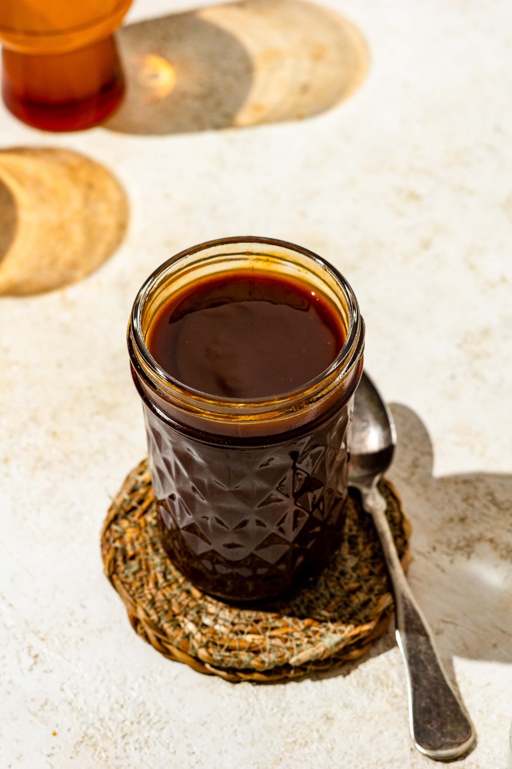 A mason jar with chicken Katsu sauce. The jar is on a tan counter on a woven coaster and a spoon.