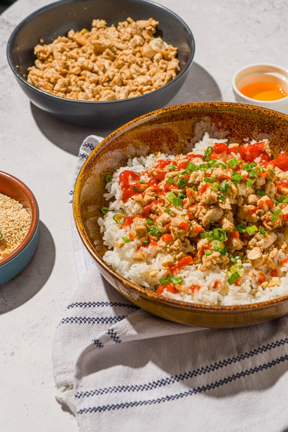 A bowl with ground turkey rice bowls with white rice, seasoned ground turkey, chopped peanuts, sliced green onions, sesame seeds, and drizzled with sriracha sauce. The bowl is on a white counter with a white striped napkin and skillet with cooked ground turkey.