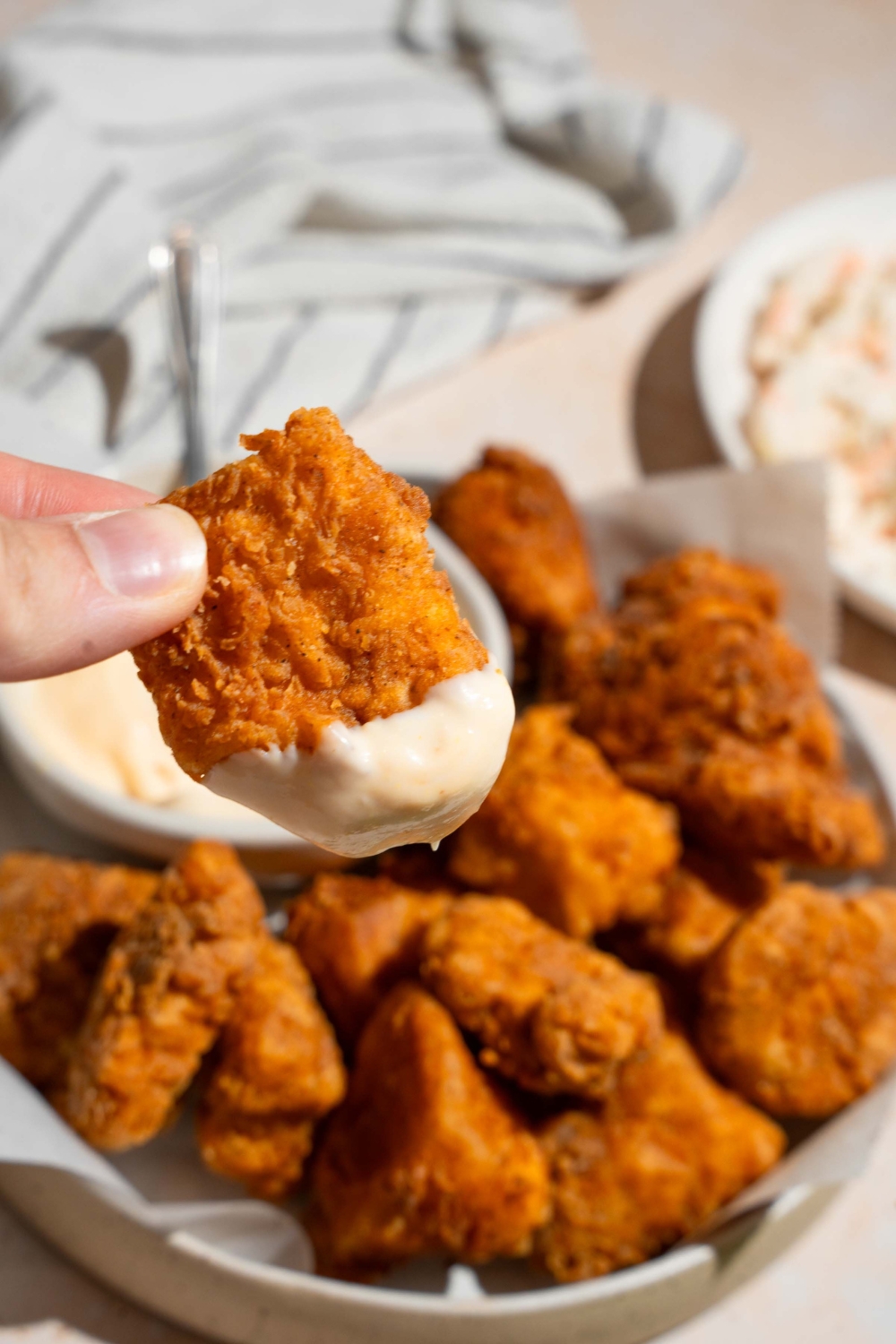 A close up of hand holding a homemade Chick Fil A nugget dipped in sauce. There is a plate of nuggets with homemade sauce blurred in the background.