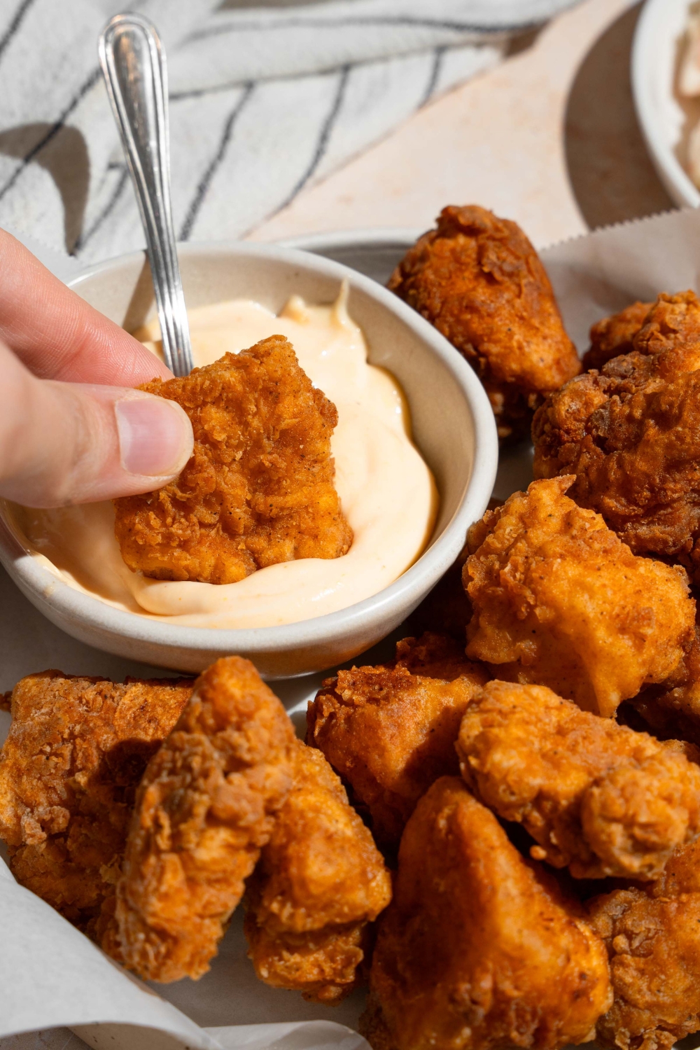 A hand dipping a homemade Chick Fil A nugget in a small bowl of sauce. The bowl is on a plate with nuggets lined with parchment paper.