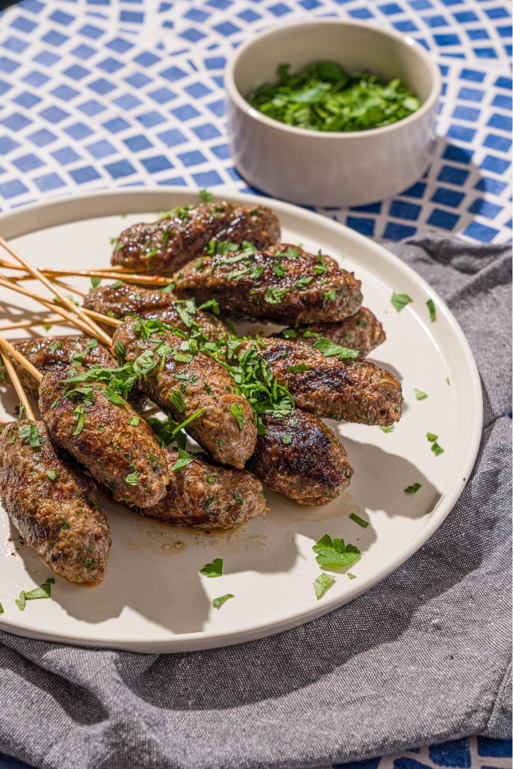 A white plate with a pile of Kafta kabobs garnished with fresh parsley. The plate is on a tiled counter with a gray cloth napkin.