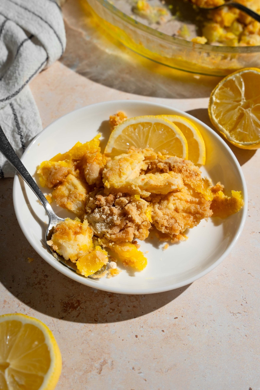 A white plate with a slice of lemon cream cheese dump cake served with sliced lemon and a fork. The plate is on a tan counter with a baking dish with cake, lemons, and a white striped napkin.