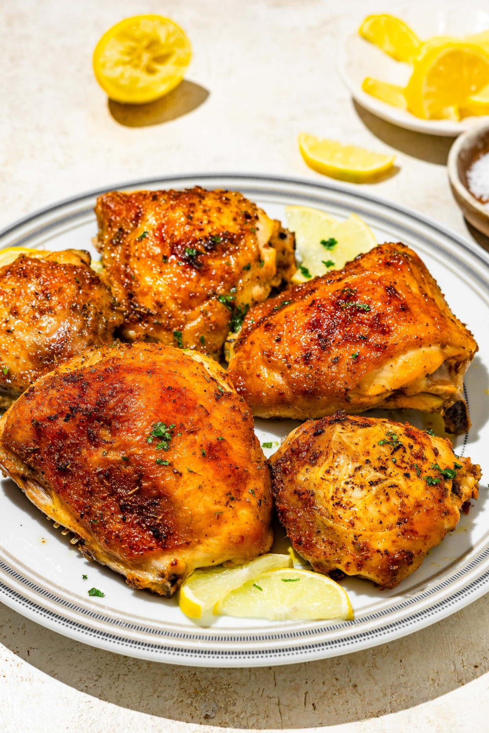 A plate of lemon pepper chicken thighs garnished with fresh parsley and lemon. The plate is on a tan counter with lemon.