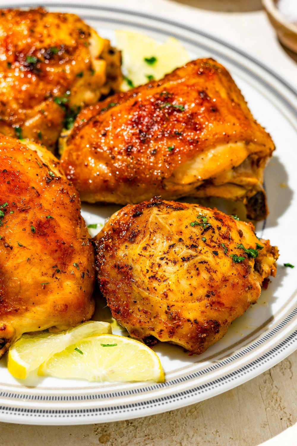 A plate of lemon pepper chicken thighs garnished with fresh parsley and lemon. The plate is on a tan counter with lemon.