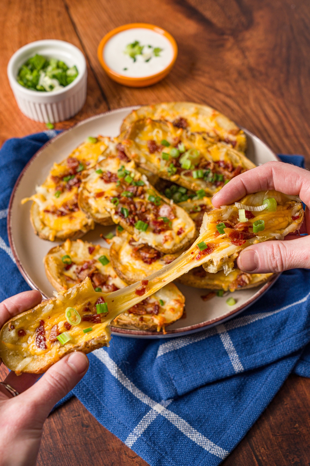 A close up of a hand pulling apart a loaded potato skin with the cheese attached. There is a plate of loaded potato skins garnished with sliced green onion. The plate is on a wood counter with a blue striped napkin.