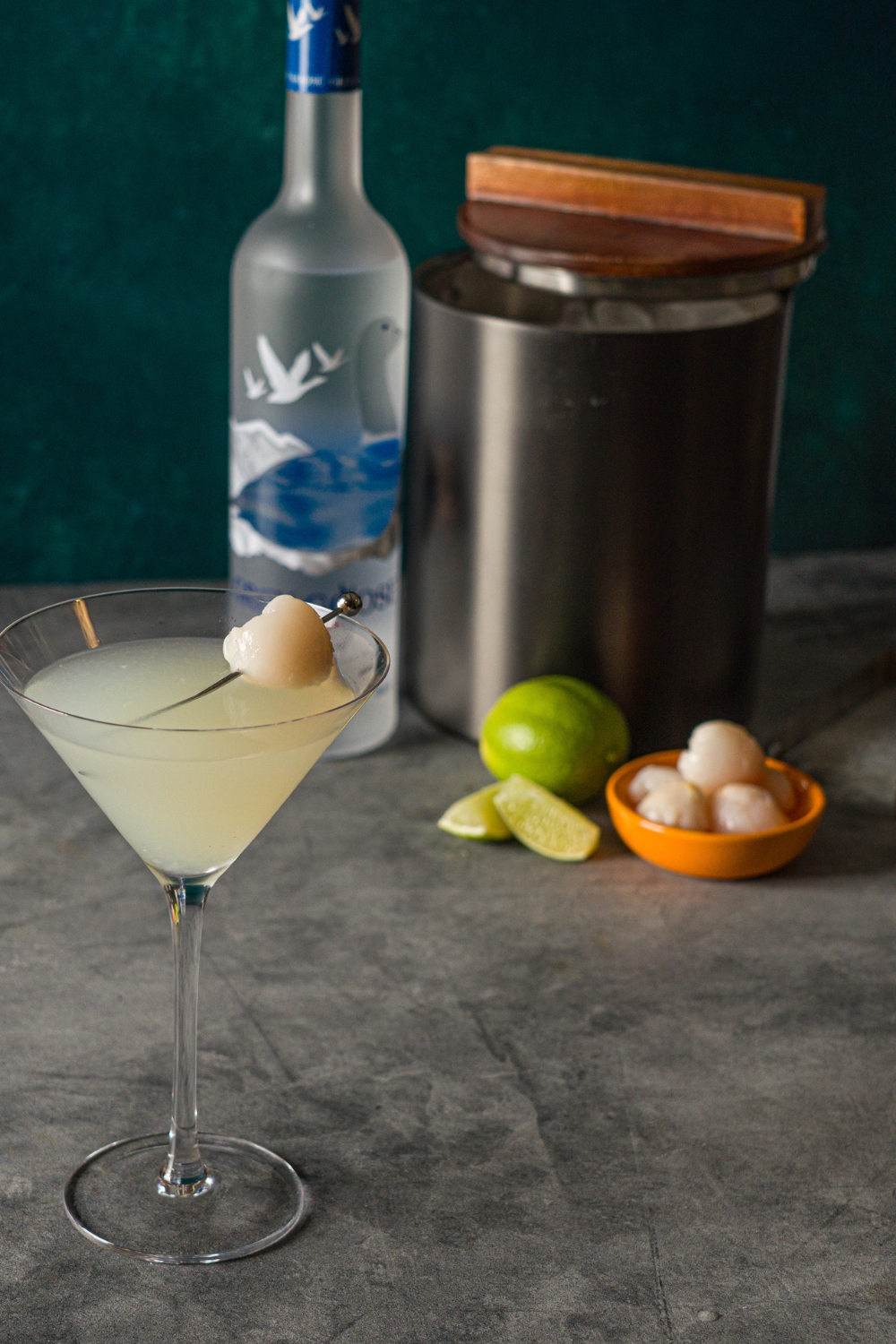 A lychee martini garnished with a lychee with a toothpick. The martini is on a stone counter with a bottle of Grey Goose, limes, small bowl of lychees, and ice bucket.