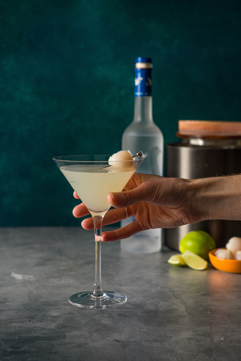 A hand holding a lychee martini garnished with a lychee with a toothpick. The martini is on a stone counter with a bottle of Grey Goose, limes, small bowl of lychees, and ice bucket.