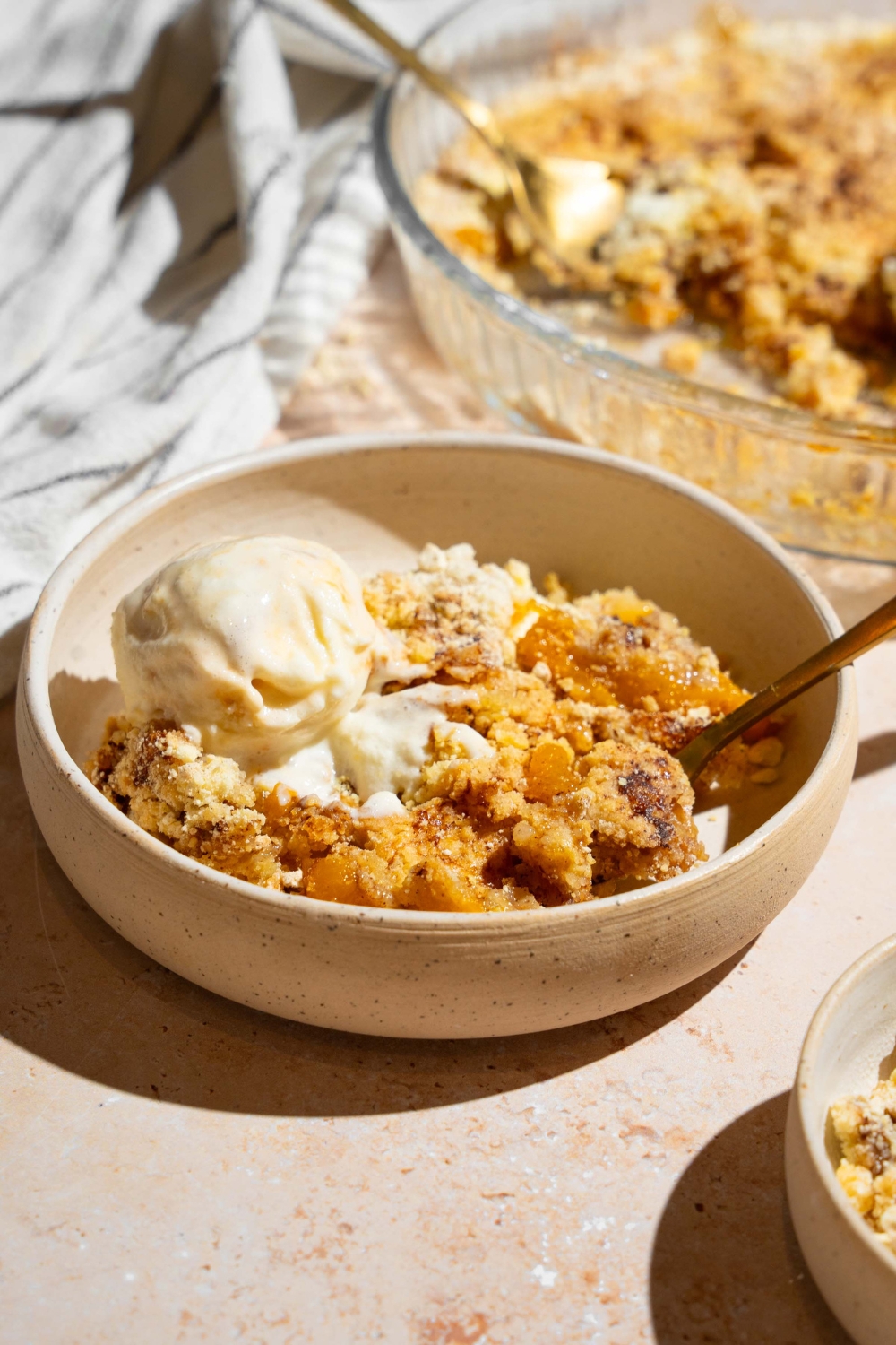 A white plate with peach cobbler dump cake topped with vanilla ice cream with a spoon. The plate is on a tan counter with a baking dish of cake.