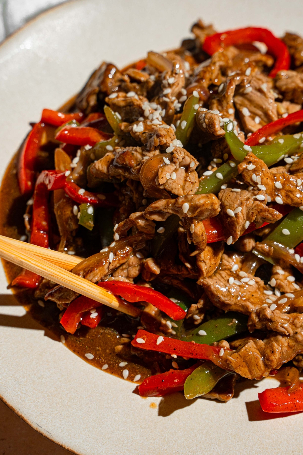 A white plate with pepper steak garnished with sesame seeds. A pair of chopsticks is taking a bite of steak.