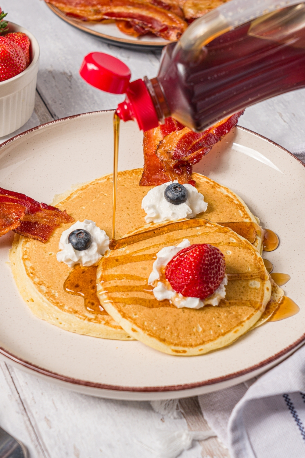 A white plate with reindeer pancakes topped with whipped cream, blueberry eyes, and a strawberry nose with bacon strips as antlers. There is pancake syrup being poured on the pancakes. The plate is on a wood counter with a bowl of berries.