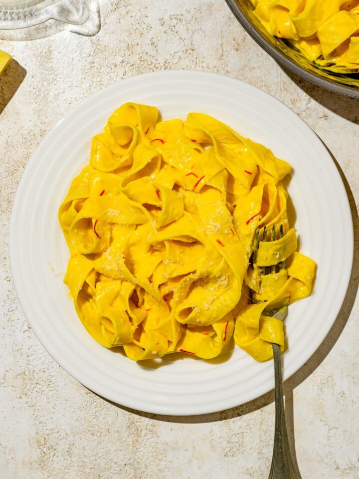 A white plate with fettuccine pasta tossed in saffron cream sauce. A fork has a bite of pasta twirled around it. The plate is on a tan counter with a skillet of pasta.