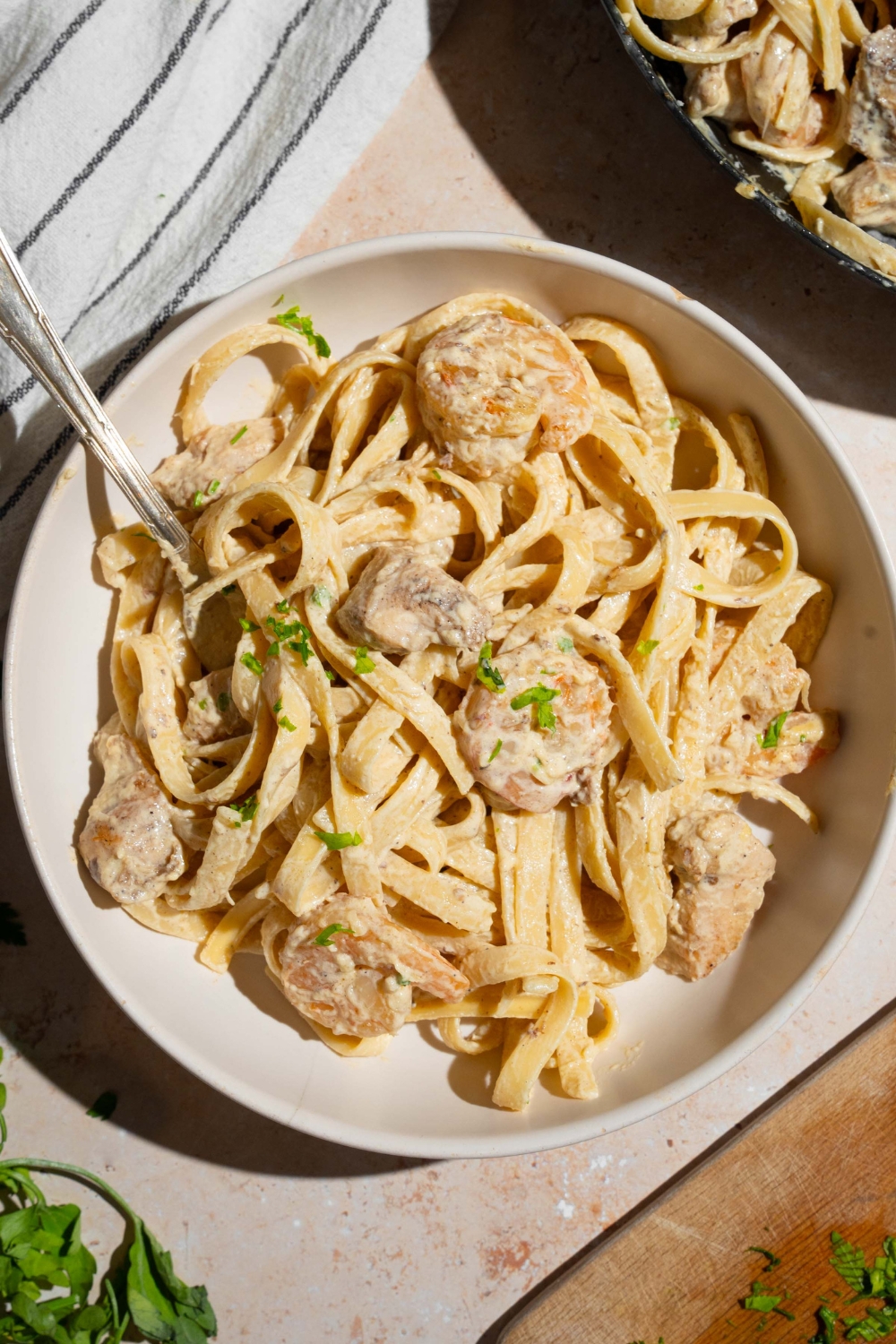 A bowl of chicken and shrimp alfredo garnished with fresh parsley. There is a fork in the bowl. The bowl is on a tan counter with white striped napkin and additional skillet of alfredo.