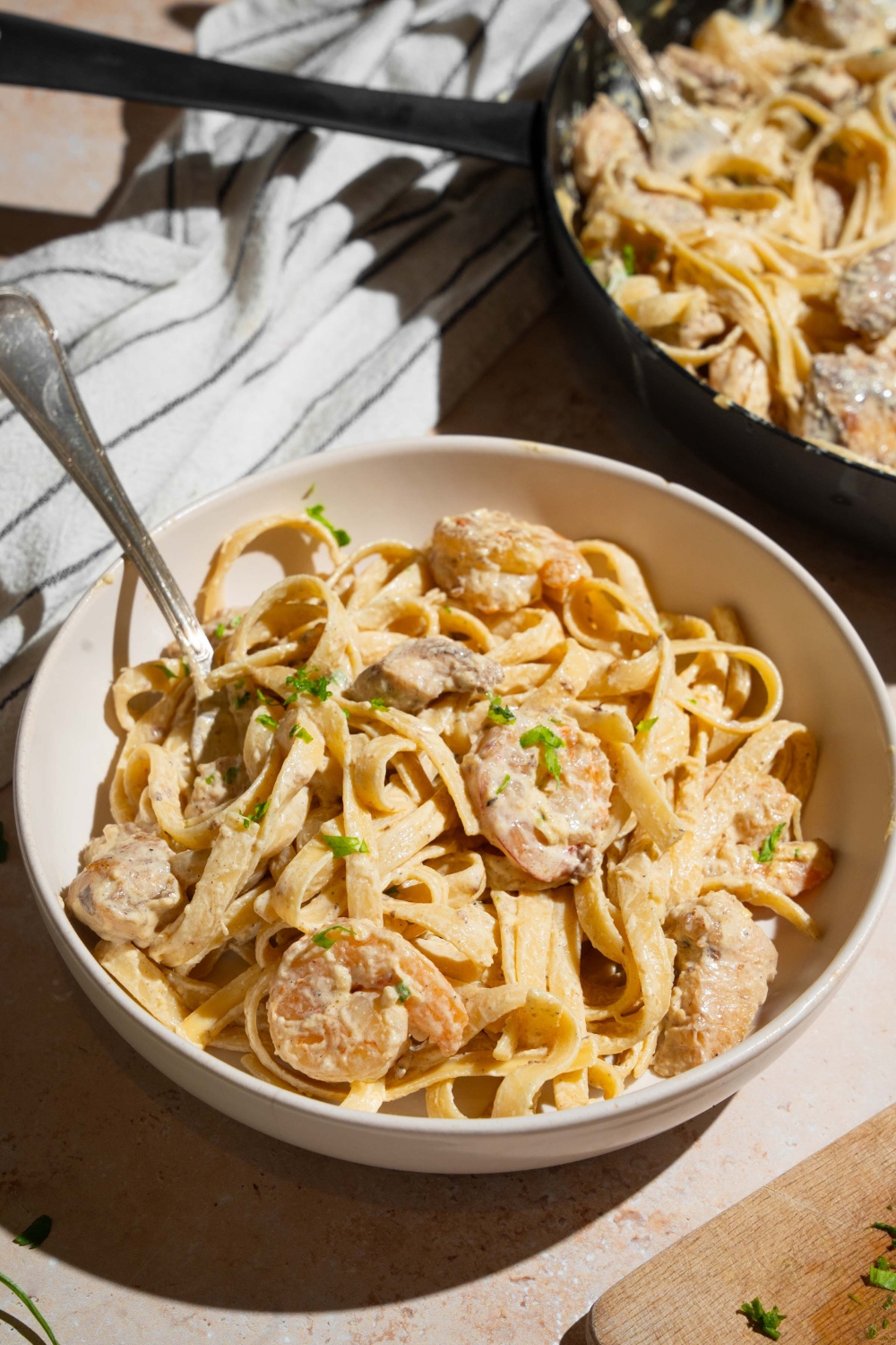 A bowl of chicken and shrimp alfredo garnished with fresh parsley. There is a fork in the bowl. The bowl is on a tan counter with white striped napkin and additional skillet of alfredo.
