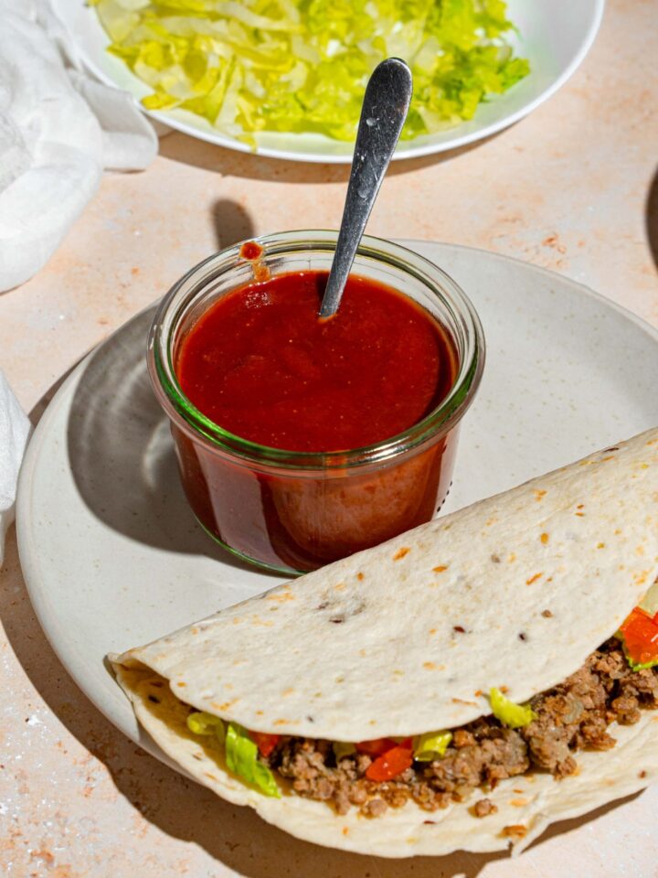 A glass jar of taco sauce with a spoon in the jar. The jar is on a white plate with a soft shell taco. The plate is on a tan counter with a plate of shredded lettuce.