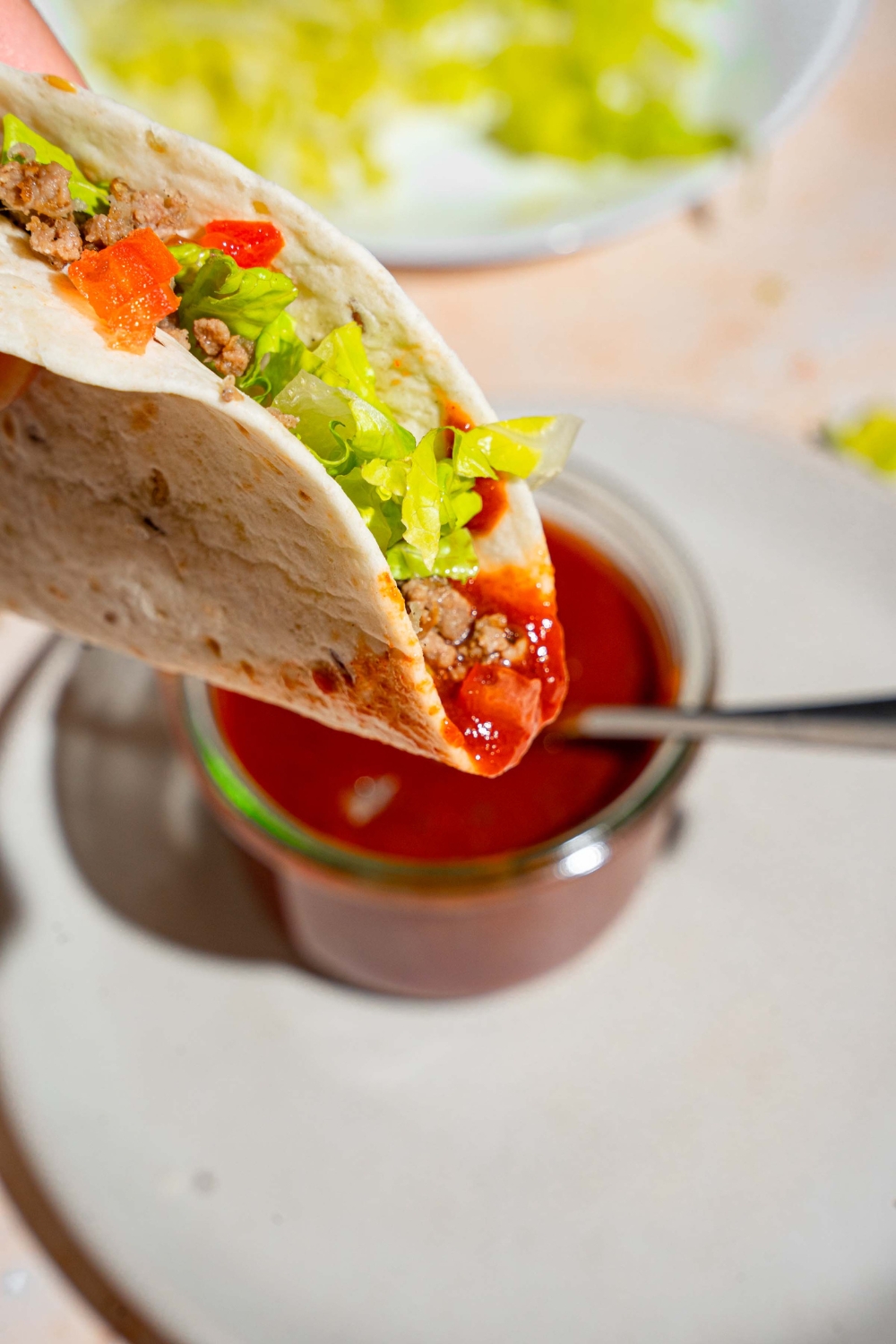 A close up of a soft shell taco dipped in taco sauce. There is a jar of taco sauce on a white plate blurred in the background.