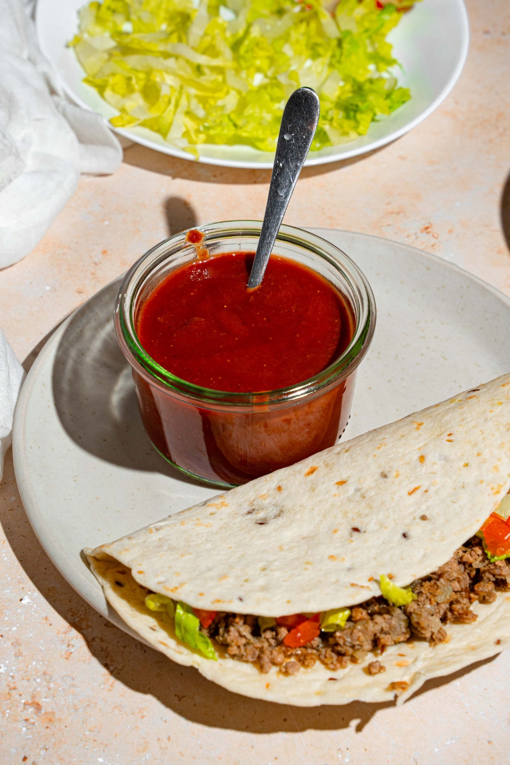 A glass jar of taco sauce with a spoon in the jar. The jar is on a white plate with a soft shell taco. The plate is on a tan counter with a plate of shredded lettuce.