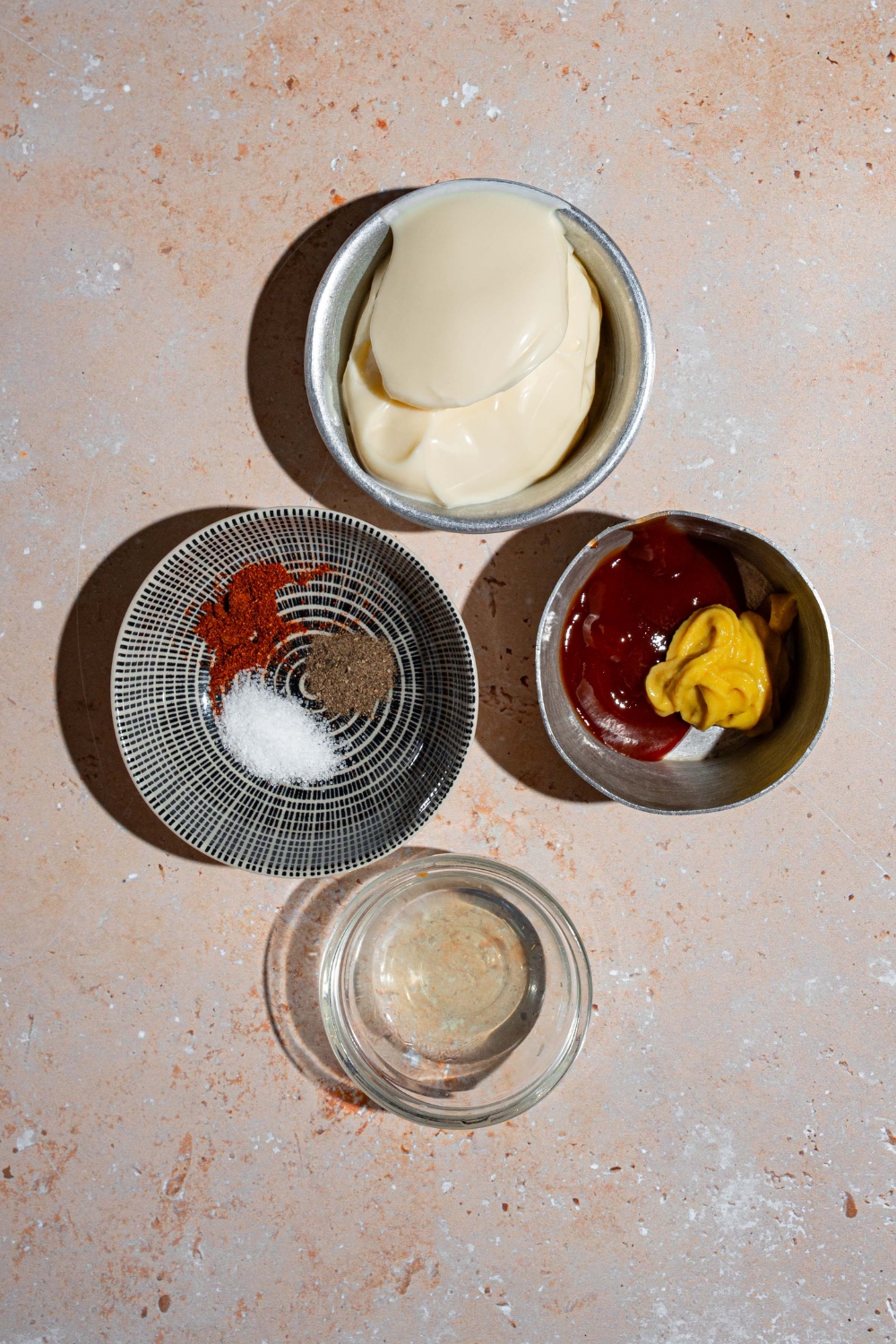 An overhead shot of several bowls in various sizes containing ingredients to make smash burger sauce including mayo, ketchup, mustard, brine, and seasonings.