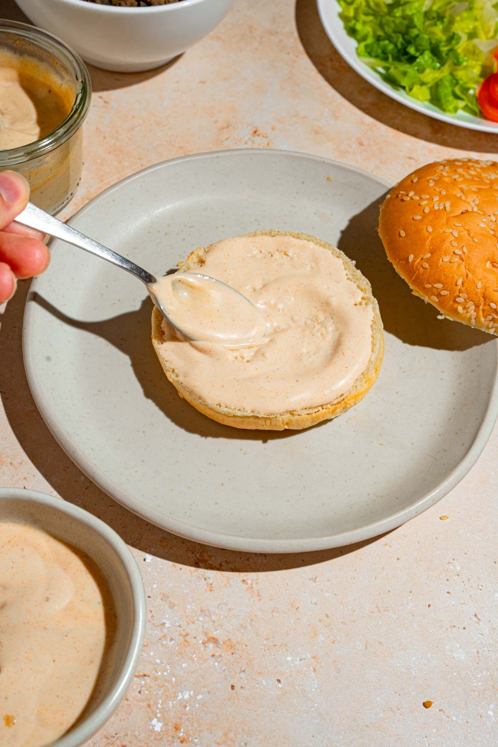 A white plate with a spoon spreading smash burger sauce on the bottom half of a burger bun. The plate is on a tan counter with additional bowls of sauce.