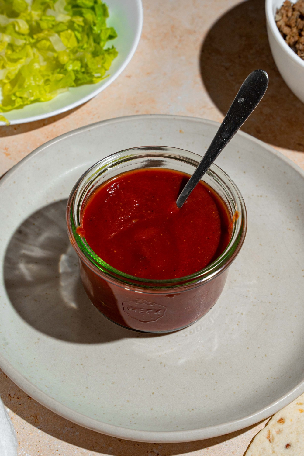 A glass jar of taco sauce with a spoon in the jar. The jar is on a white plate. The plate is on a tan counter with a plate of shredded lettuce.