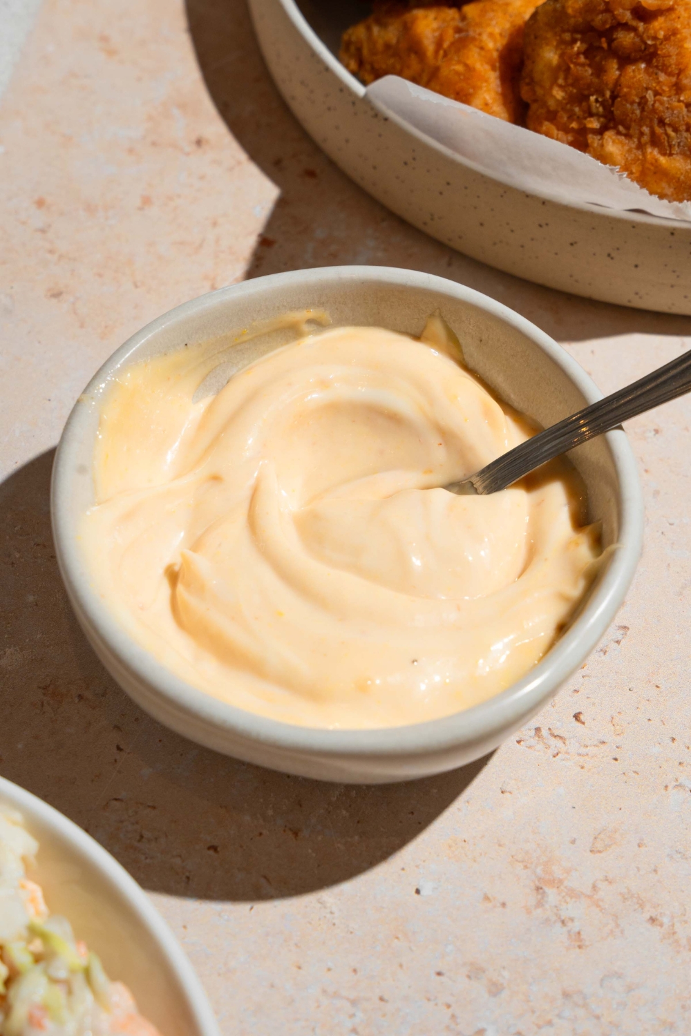 A small bowl with a spoon mixing homemade Chick Fil A sauce. The bowl is on a tan counter with a plate of nuggets.