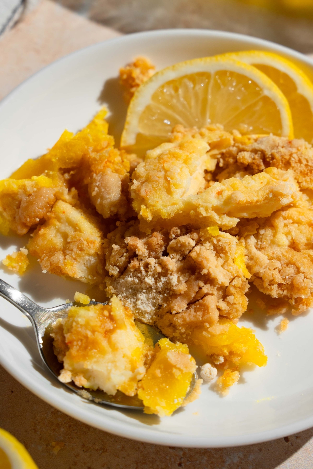 A white plate with a slice of lemon cream cheese dump cake served with sliced lemon and a fork. The plate is on a tan counter.