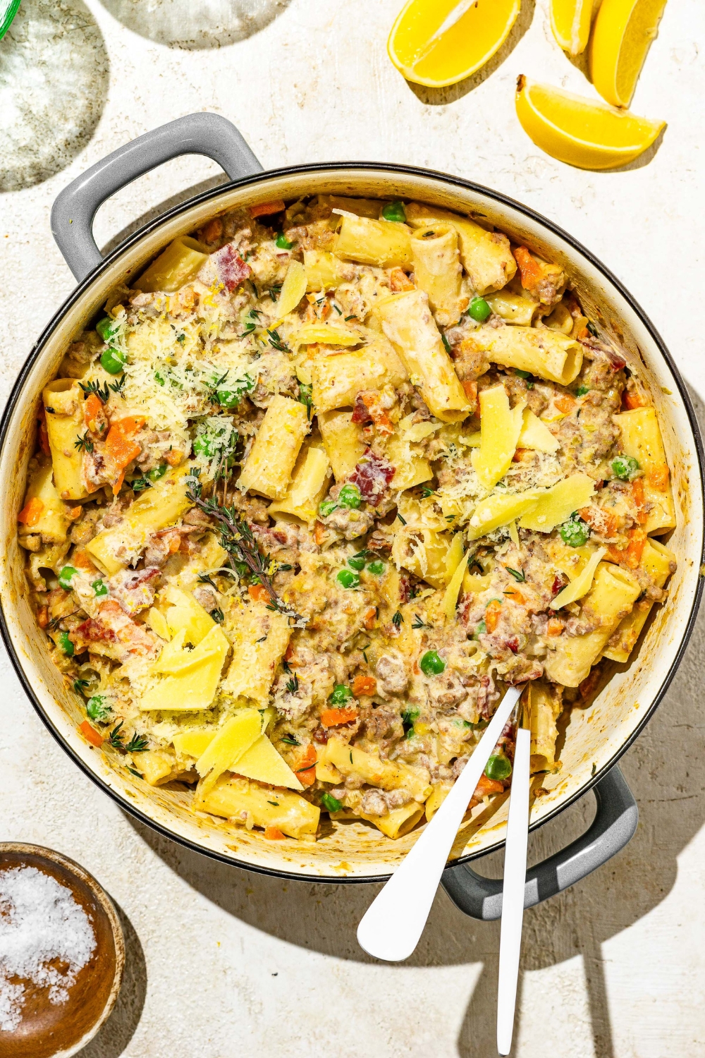 A dutch oven with rigatoni pasta with white bolognese. There is a fork and spoon in the pot. The dish is garnished with grated cheese. The pot is on a tan counter with sliced lemon and a small bowl of garnishes.