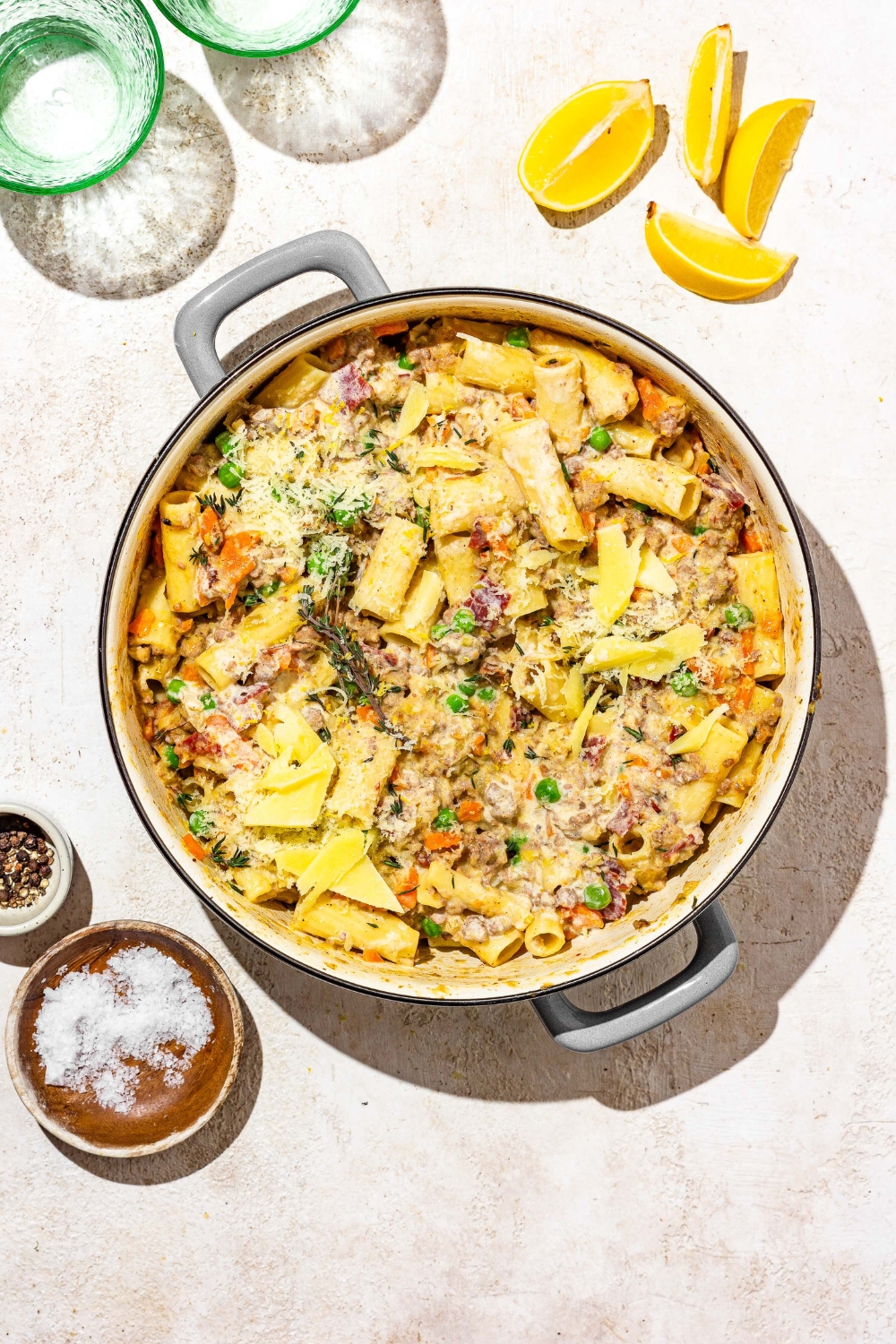 A dutch oven with rigatoni pasta with white bolognese. The dish is garnished with grated cheese. The pot is on a tan counter with sliced lemon and a small bowl of garnishes.