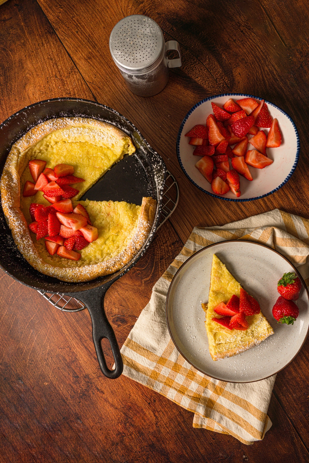 A german pancake in a skillet. A slice is out of it and on a plate next to the skillet. In front of that is a bowl of sliced strawberries.