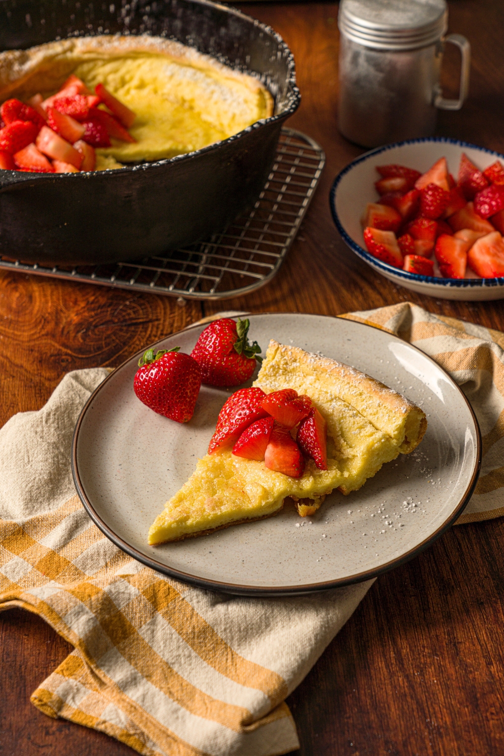 A slice of a dutch baby on a white plate. Behind it is a skillet filled with the dutch baby.