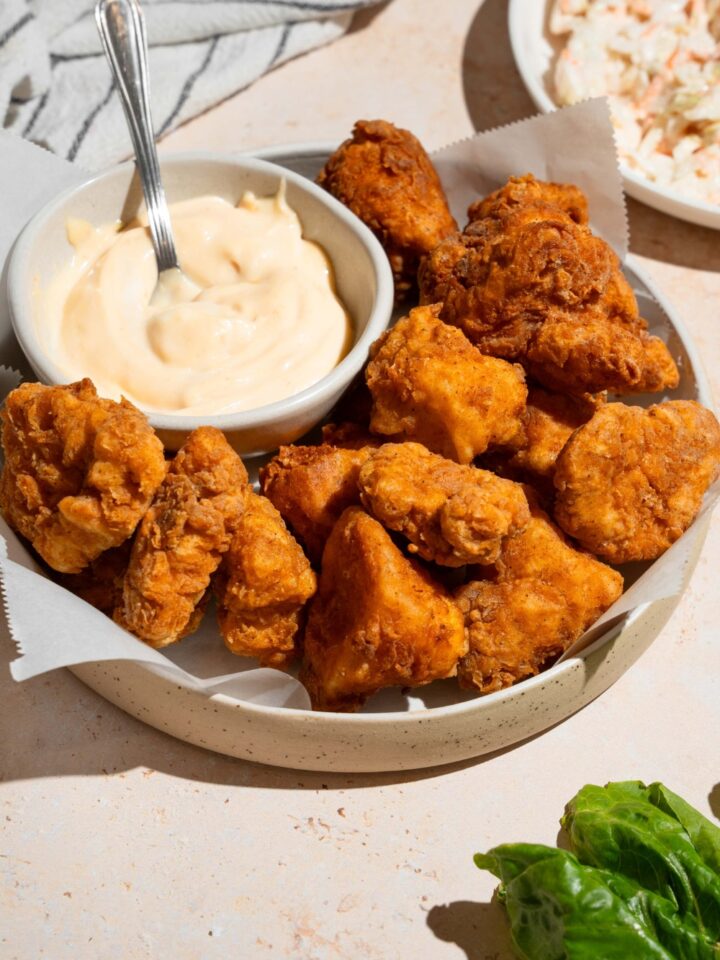 Homemade Chick Fil A nuggets and sauce on a white plate lined with parchment paper. The plate is on a tan counter with a white striped napkin and small plate of coleslaw.