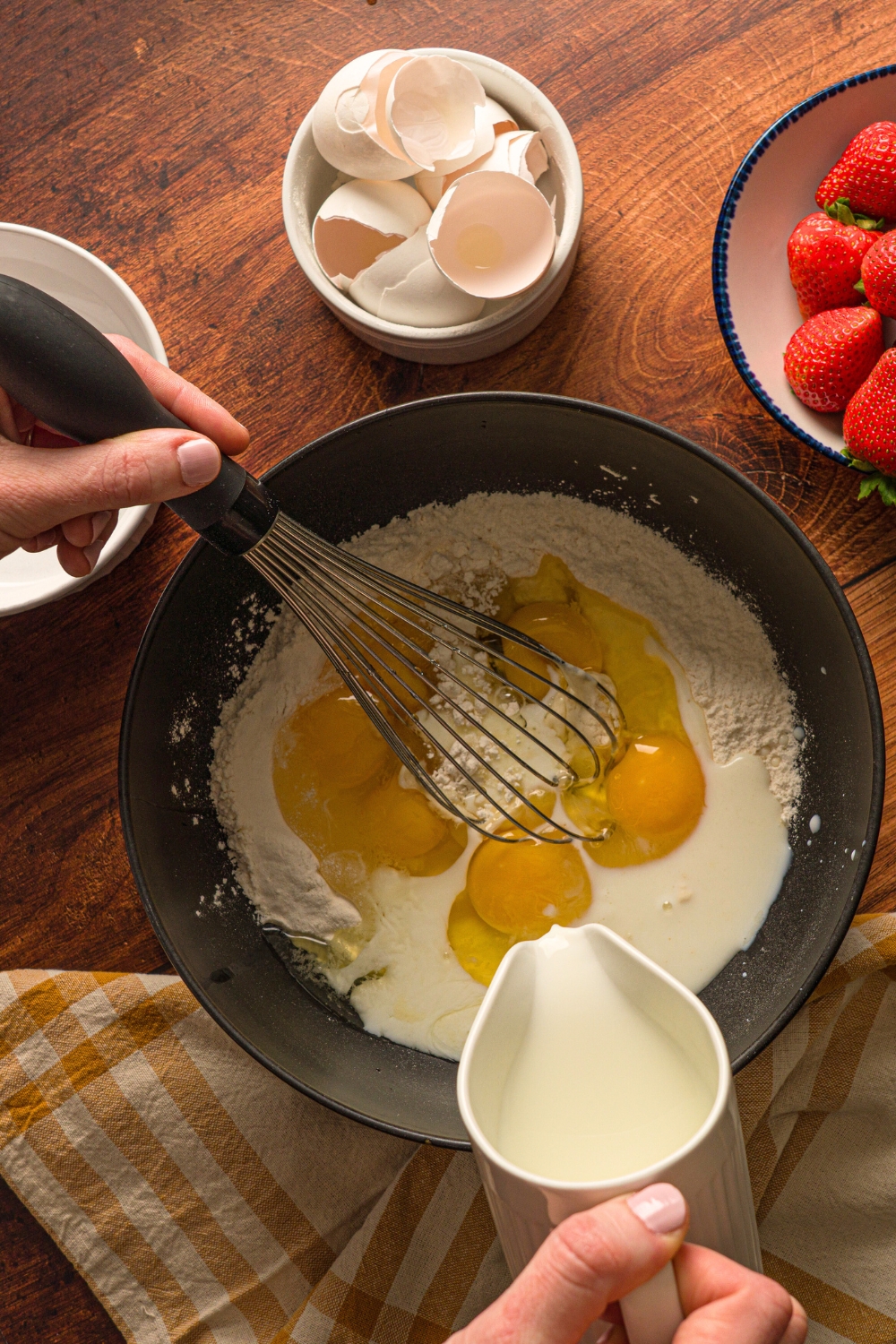 A whisk in a pot with eggs, flour, and milk being poured into it.