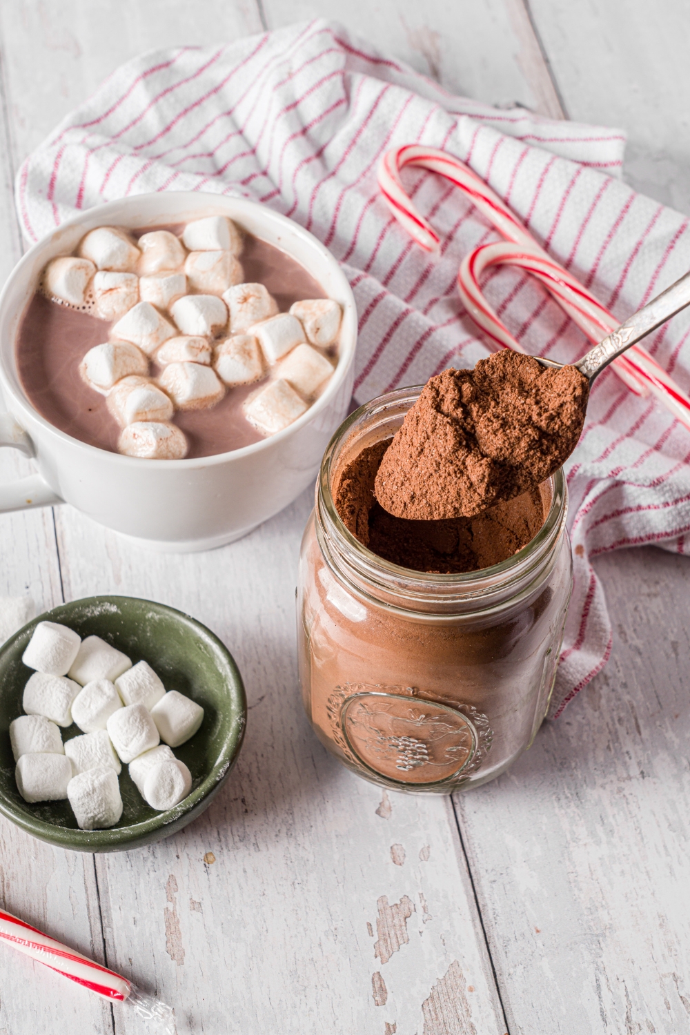 A mason jar of hot cocoa mix with a spoon scooping cocoa mix from the jar. The jar is on a wooden counter with a mug of hot cocoa topped with marshmallows, a small bowl of marshmallows, and candy canes.