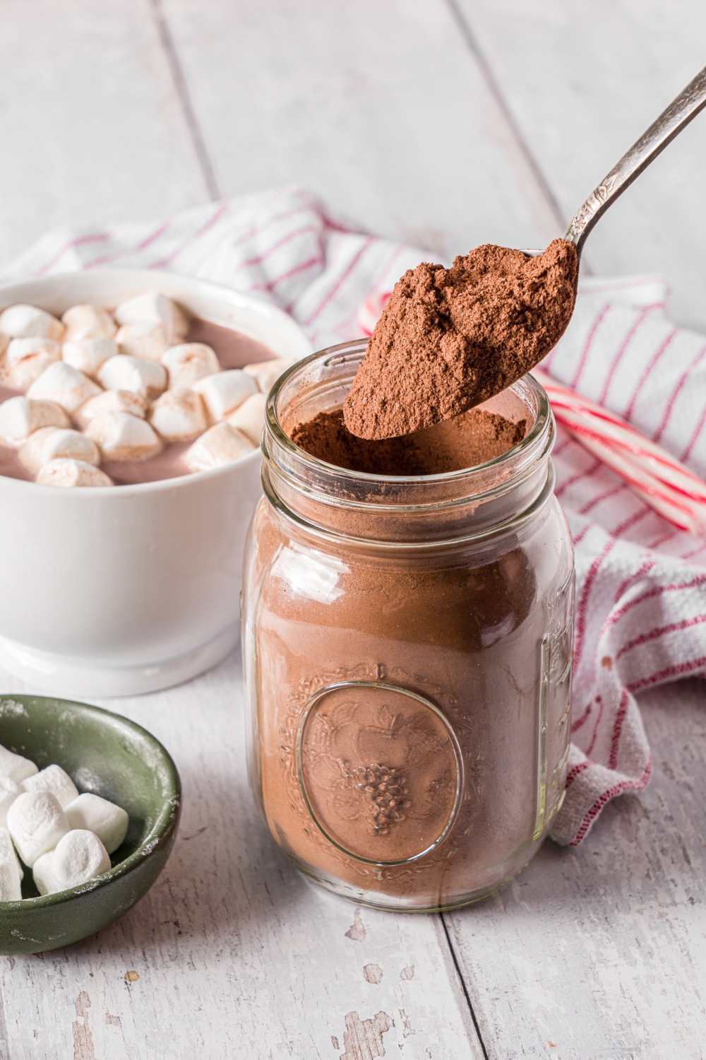 A mason jar of hot cocoa mix with a spoon scooping cocoa mix from the jar. The jar is on a wooden counter with a mug of hot cocoa topped with marshmallows, a small bowl of marshmallows, and candy canes.