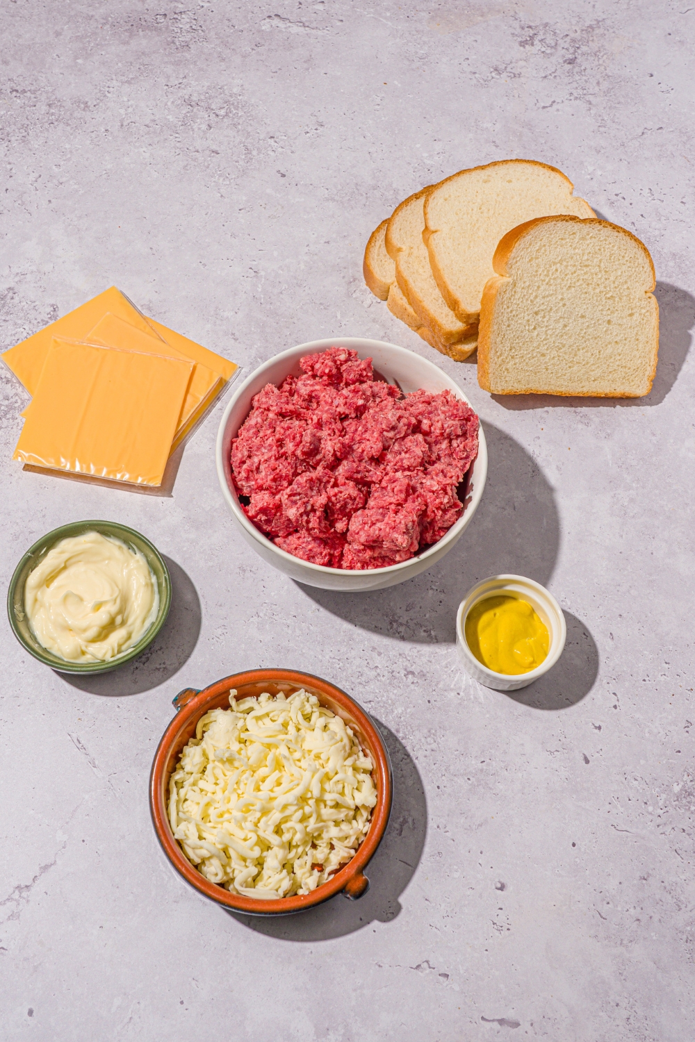 An overhead shot of ingredients to make Applebee's grilled cheeseburger including white bread, ground beef, American cheese slices, shredded mozzarella, mustard, mayo, and seasonings.