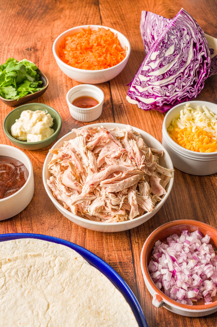 Several bowls in various sizes containing ingredients to make BBQ chicken wraps including shredded chicken, tortilla wraps, shredded cheese, red onion, purple cabbage, shredded carrots, and more.