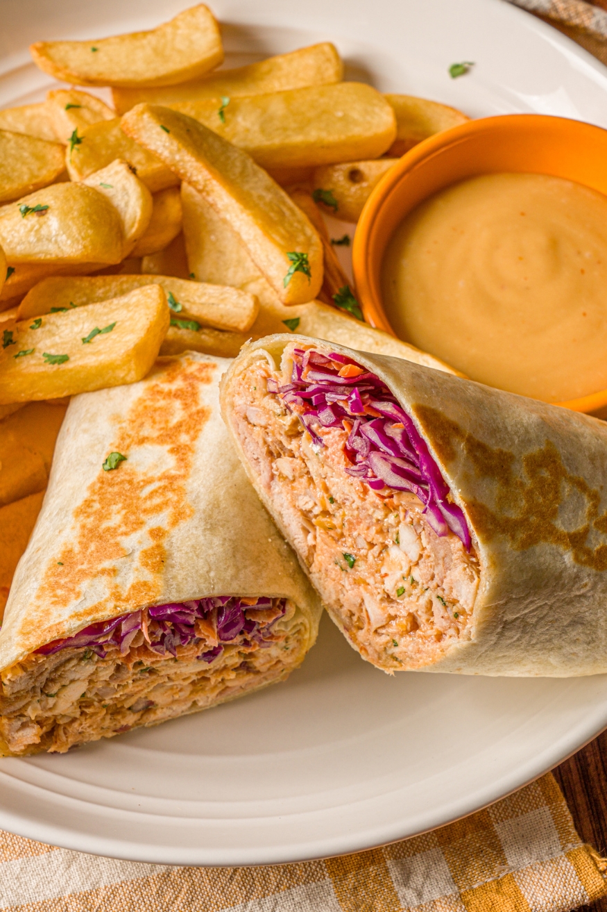 A BBQ chicken wrap sliced in half on a white plate with french fries garnished with fresh parsley. The wrap is served with a small bowl of dipping sauce.