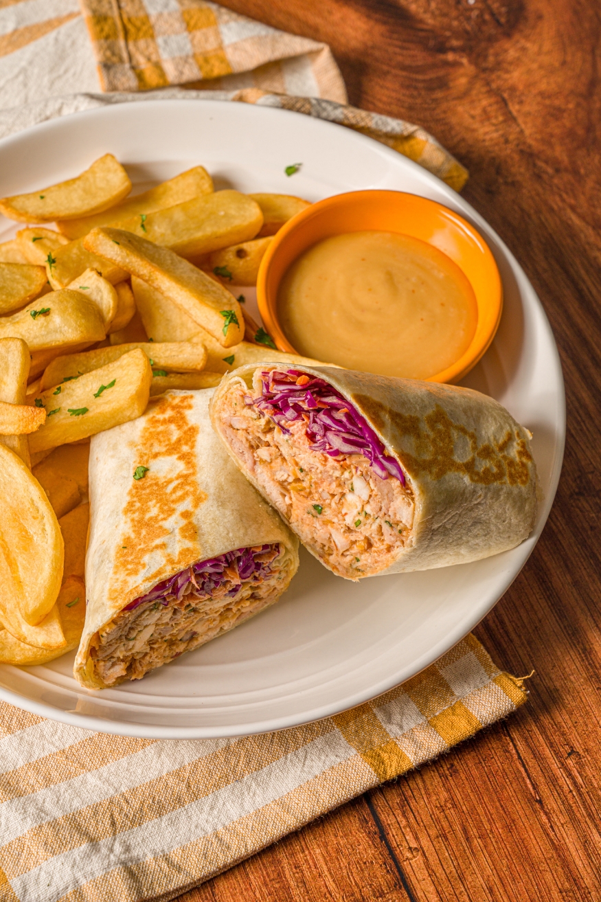A BBQ chicken wrap sliced in half on a white plate with french fries garnished with fresh parsley. The wrap is served with a small bowl of dipping sauce. The plate is on a wood counter with a yellow striped napkin.