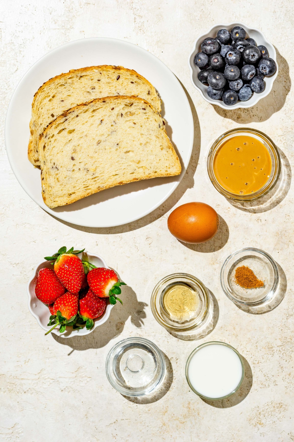 An overhead shot of several bowls in various sizes containing ingredients to make baby french toast including slices of rye bread, peanut butter, egg, cinnamon, milk, strawberries, vanilla extract, and coconut oil.