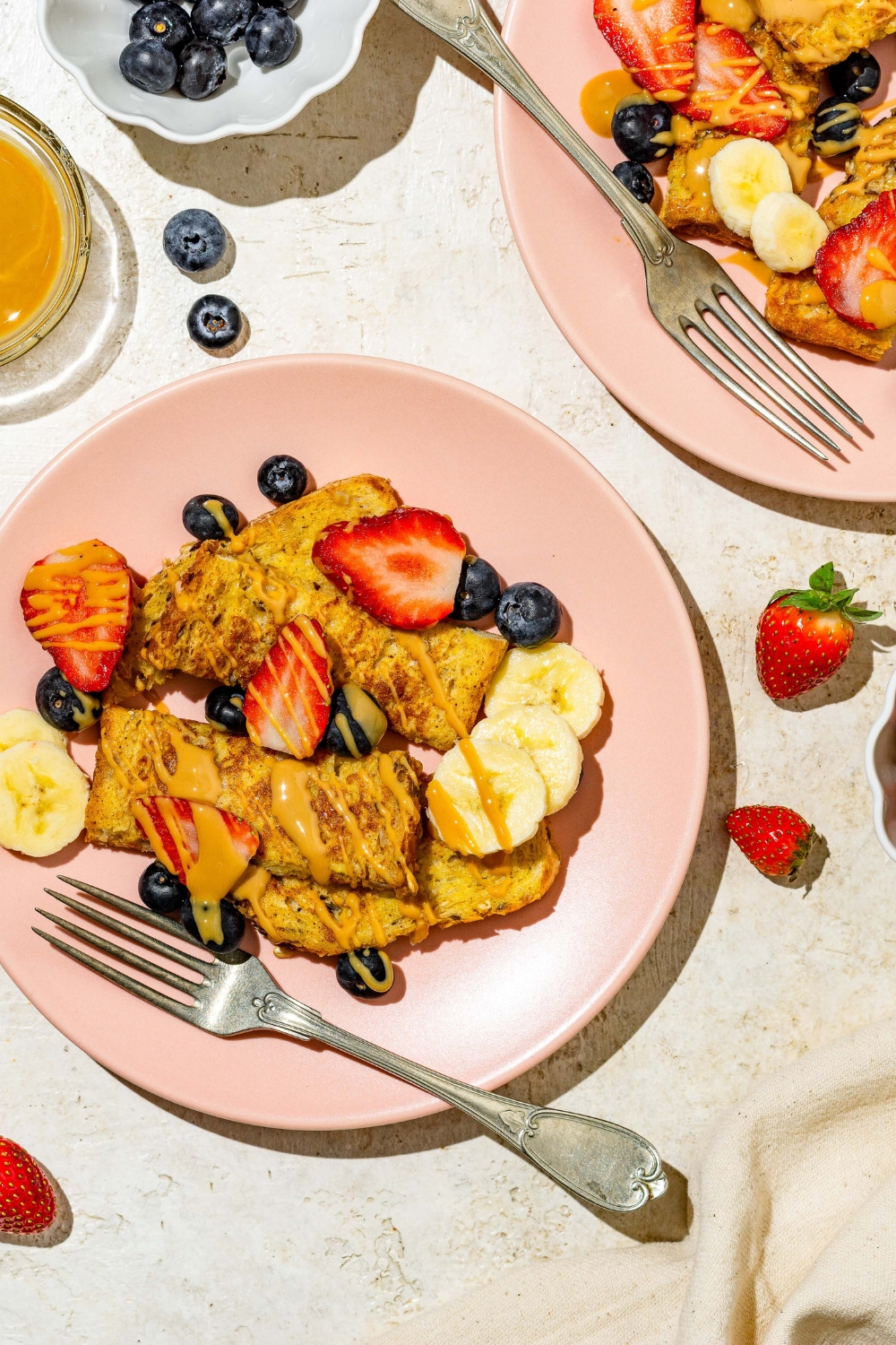 A pink plate with sliced baby french toast sticks toped with sliced banana, blueberries, and strawberries and drizzled with peanut butter. There is a fork on the plate. The plate is on a tan counter with an additional plate of french toast and small bowl of blueberries.