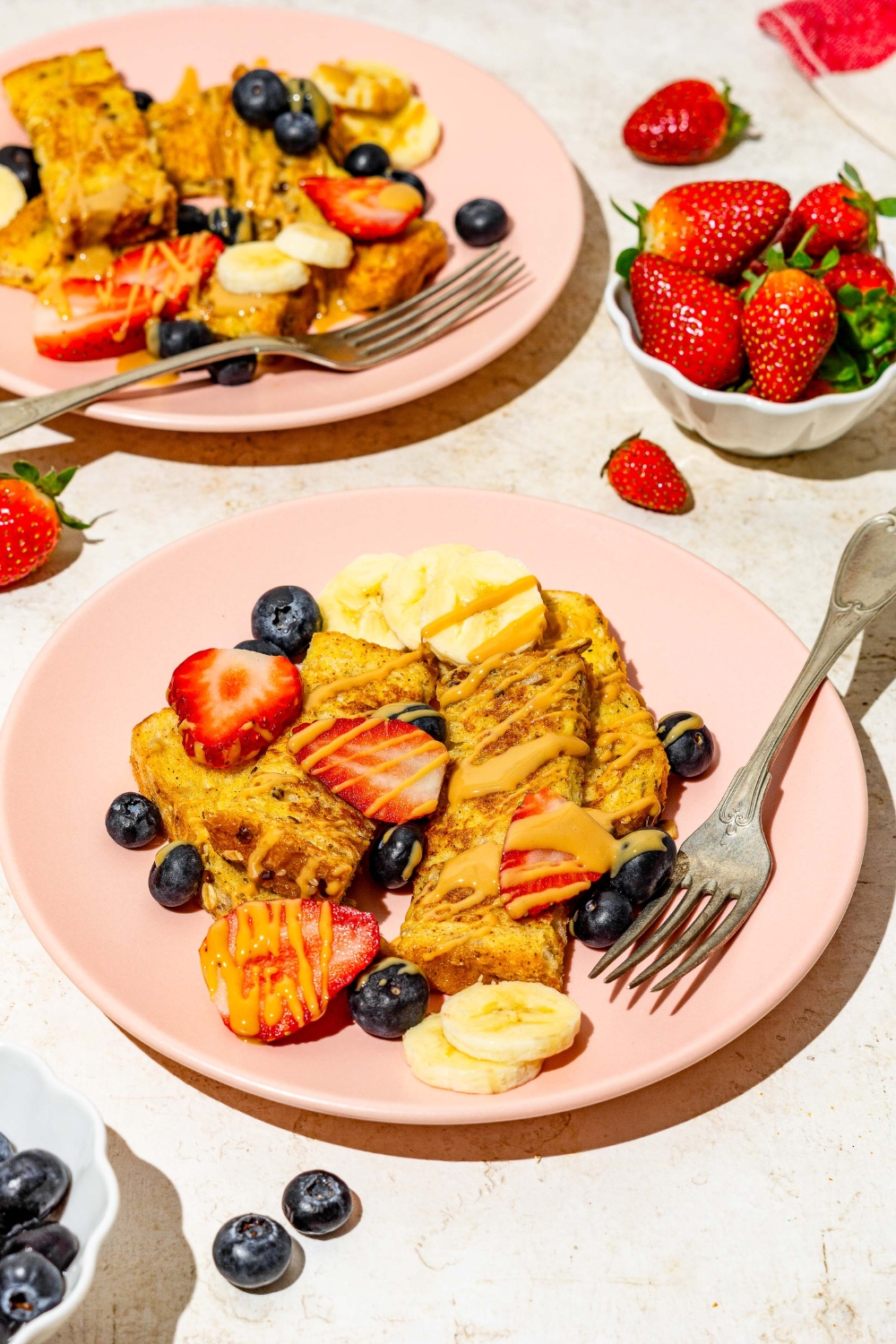 A pink plate with sliced baby french toast sticks toped with sliced banana, blueberries, and strawberries and drizzled with peanut butter. There is a fork on the plate. The plate is on a tan counter with an additional plate of french toast and small bowl of strawberries.